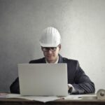 Professional engineer wearing hardhat working on construction plans at desk with laptop in office.